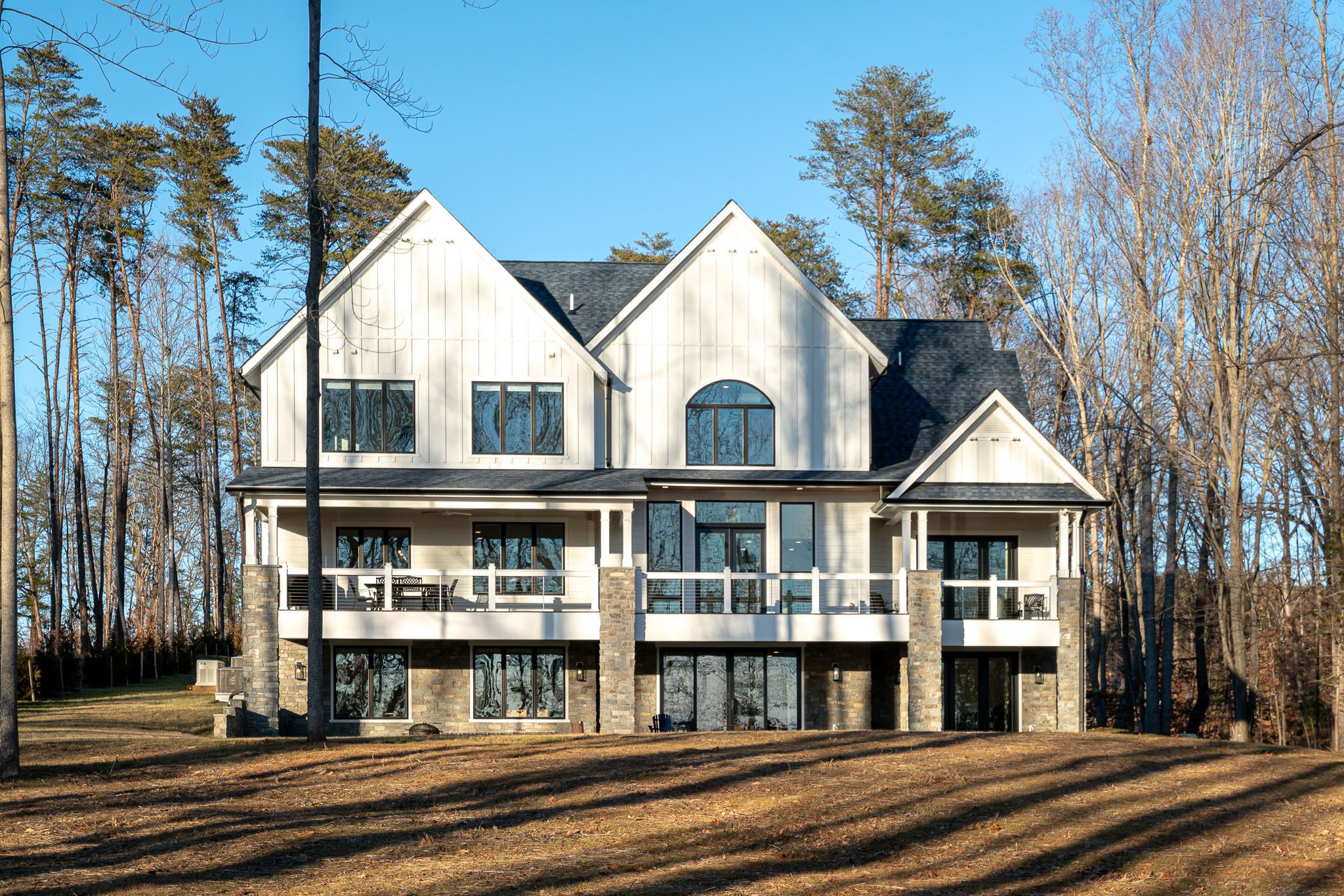 A large two-story house with white siding and numerous windows, featuring a stone foundation and surrounded by tall trees under a clear blue sky.